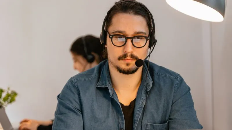 A man wearing glasses and a headset works at a desk with a colleague behind him.