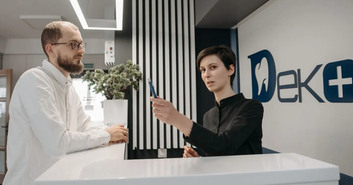A receptionist holds up a pen while talking with a man at a clinic front desk.