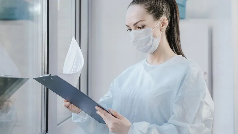 A woman in a face mask and protective gown reads papers on a clipboard.