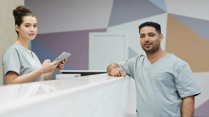 Two healthcare workers in light blue scrubs at a clinic reception desk, one holding a tablet.