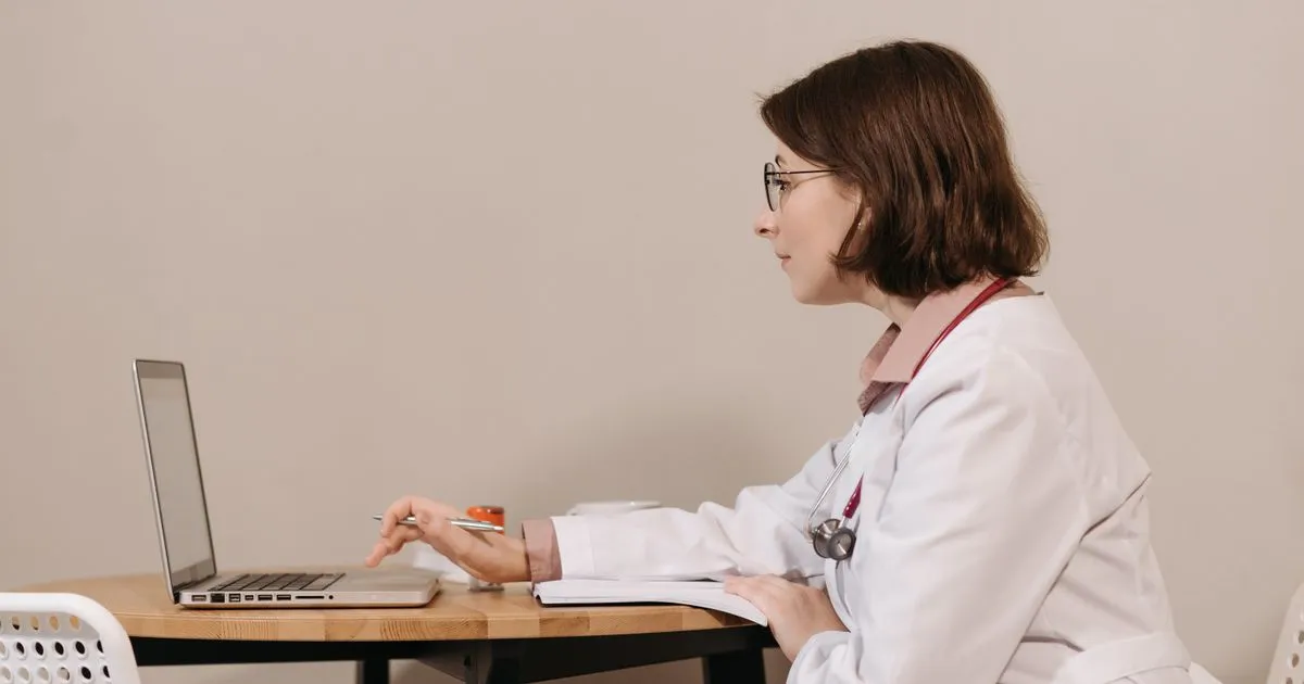 A doctor in a white coat and glasses works on a laptop at a wooden table.