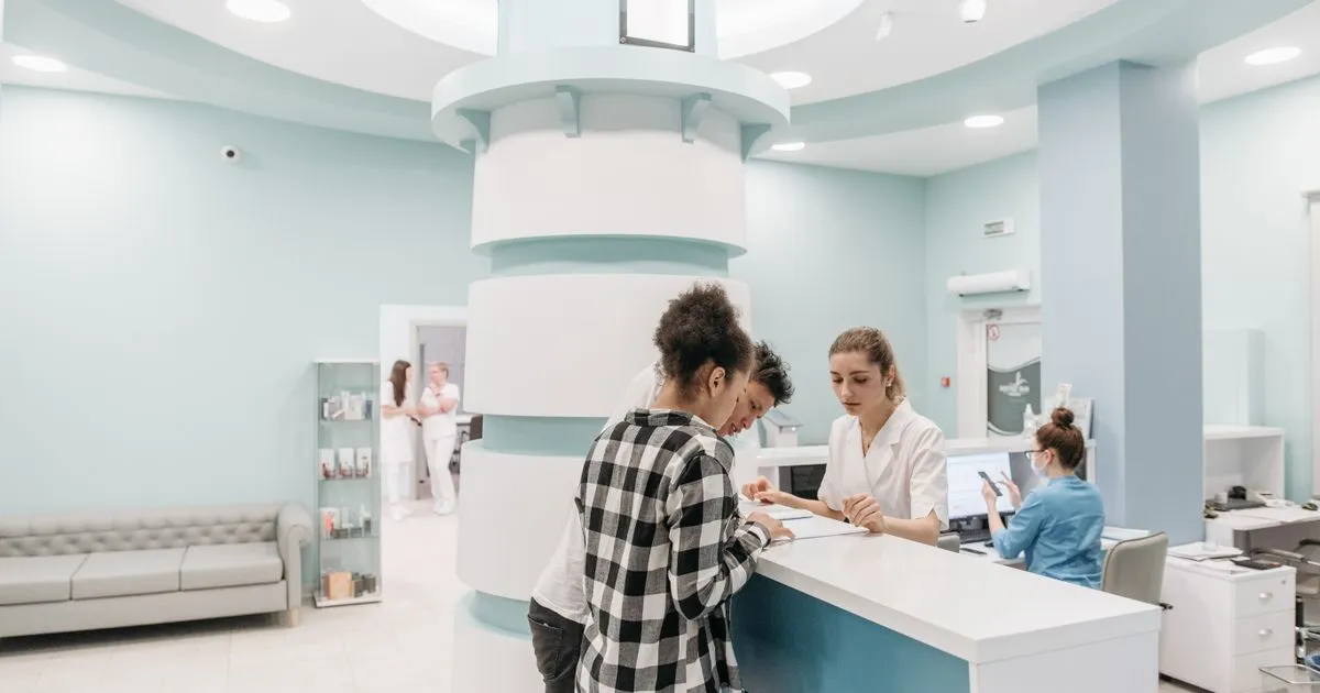 Staff member at a clinic reception desk helping two patients with paperwork.